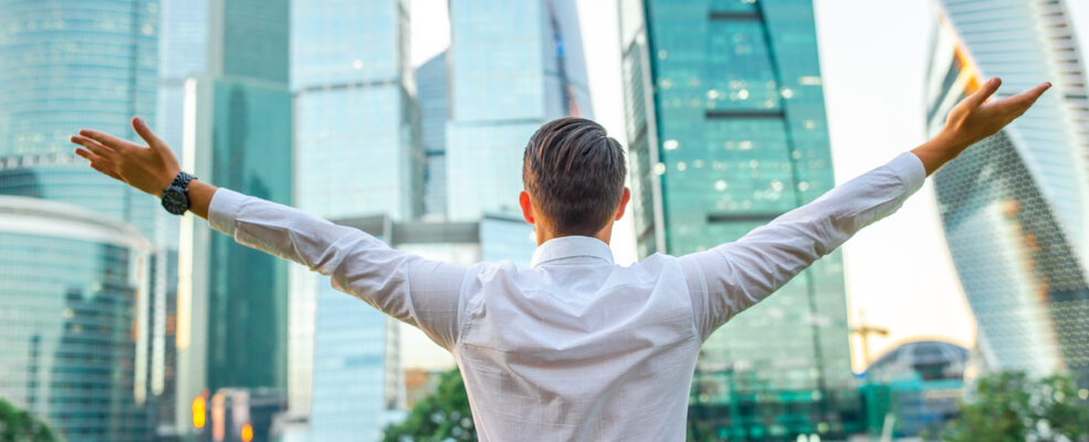 A business man stands with his arms outstretched in front of some skyscrapers. He is happy about the boundless opportunities for private engineering contracts