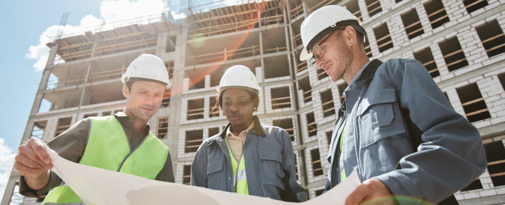 Three people in helmets looking over blueprints in front of incomplete building, they are talented engineers.