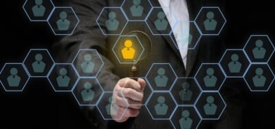 A man in a suit holds a magnifying glass over icons of business people as a metaphor for finding and recruiting talent.